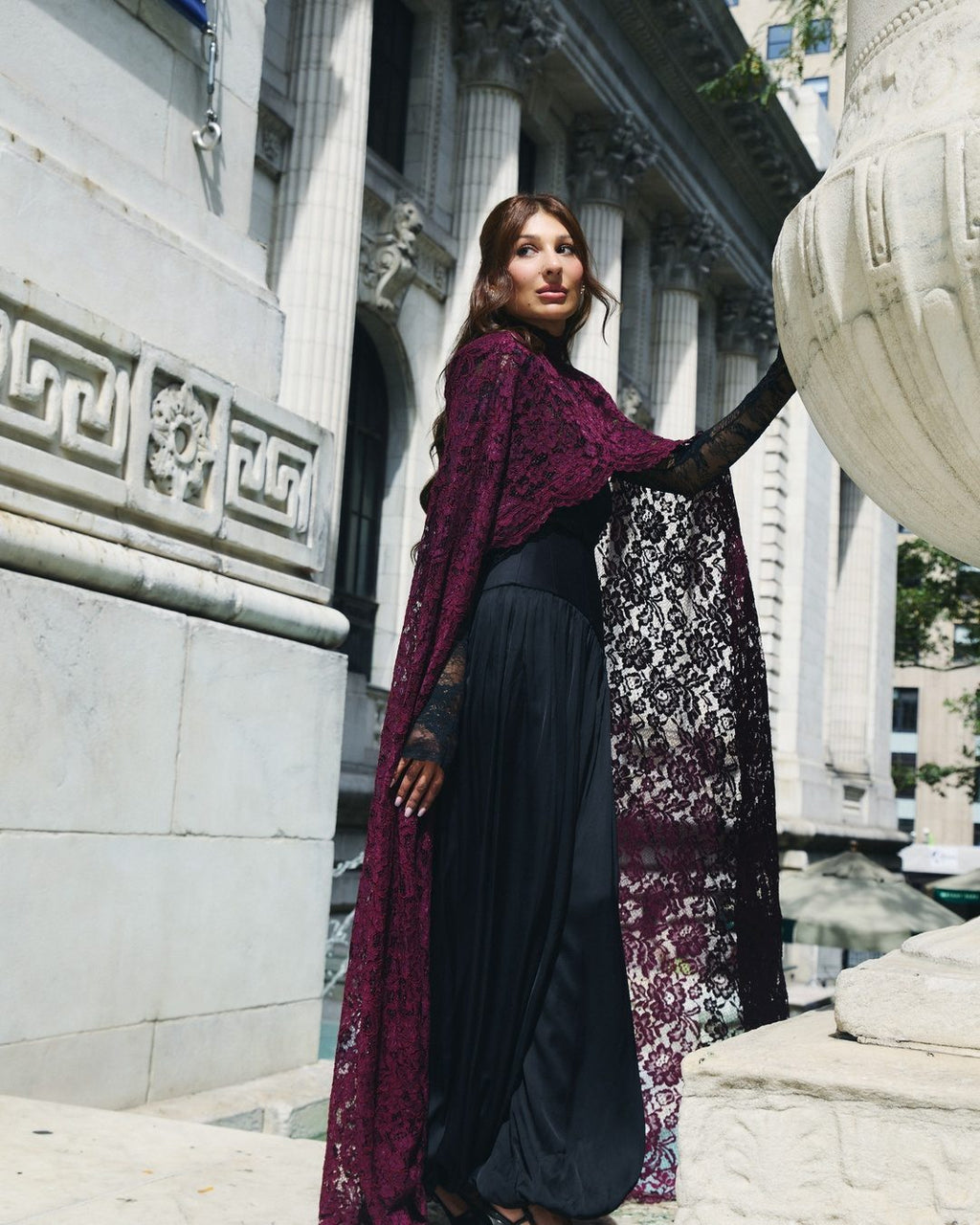 Oxblood lace cape and corset set, side view against a carved stone facade.