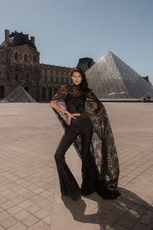 Black wide-leg trousers styled with a sheer black lace layer, the Louvre pyramid behind.